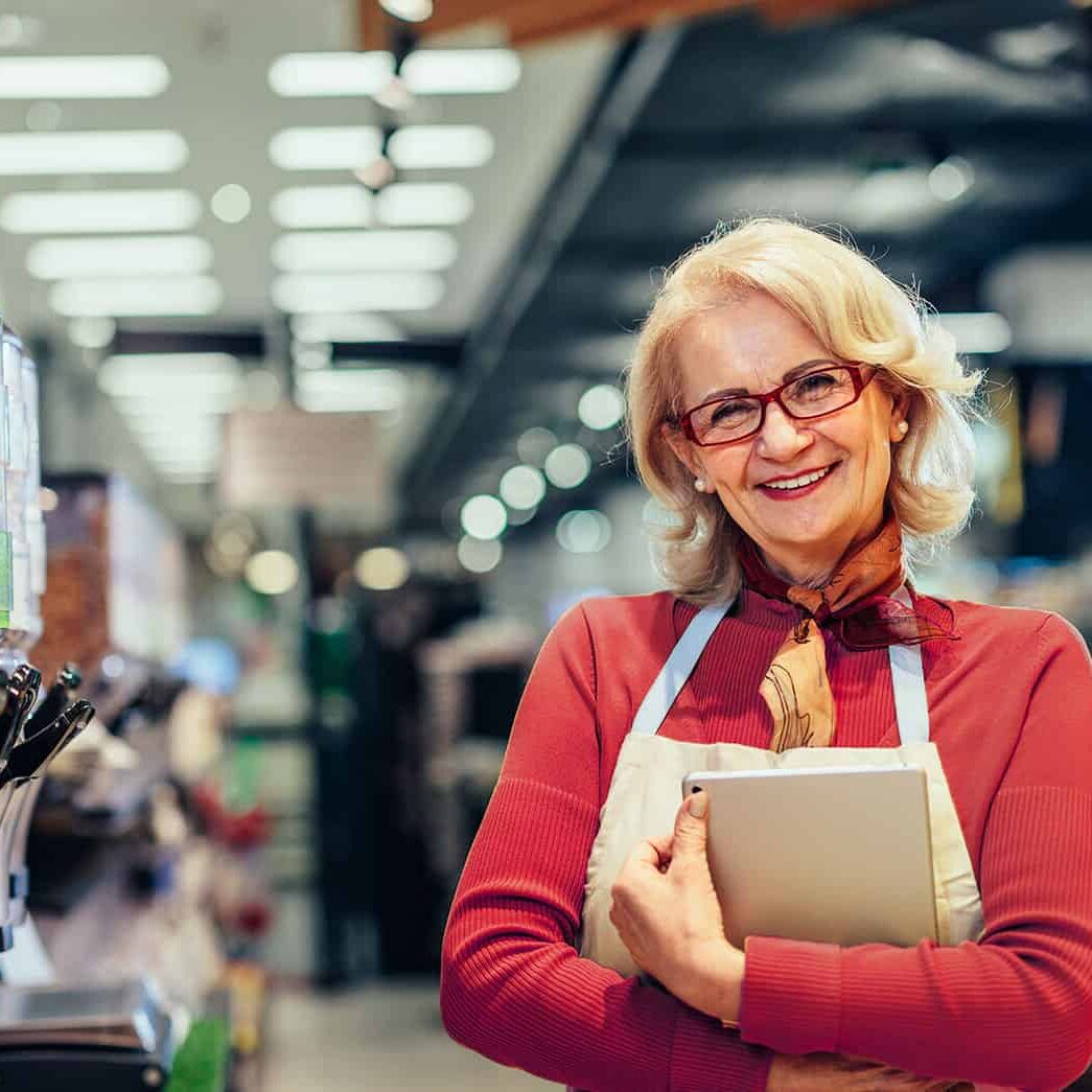Store clerk smiling in grocery store, holding a tablet and wearing an apron, showcasing inventory management, friendly customer service, and retail expertise in a supermarket environment.