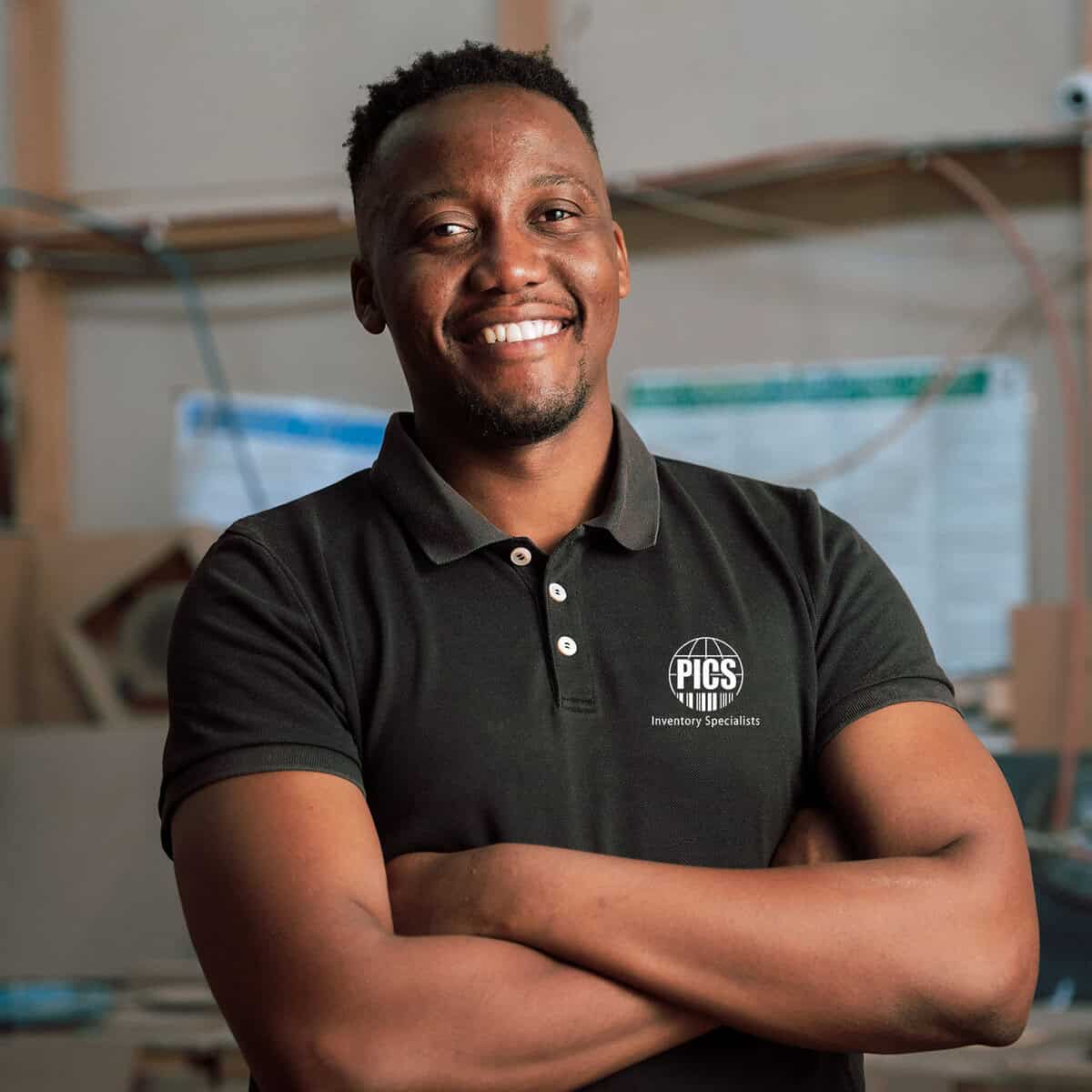 Inventory specialist smiling confidently in warehouse setting, wearing black polo shirt with PICS logo, cross arms and professional pose.