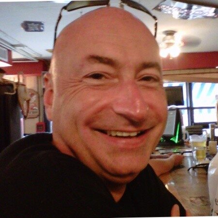 Happy man smiling in a cozy indoor setting, possibly at a restaurant or bar, with warm lighting and casual ambiance.